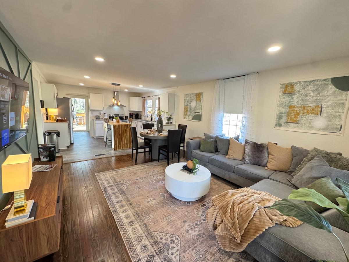 The inviting living area features a spacious sectional sofa adorned with assorted cushions, complemented by a round coffee table. An area rug anchors the space, while natural light pours in through the windows, illuminating the adjacent dining area and modern kitchen.