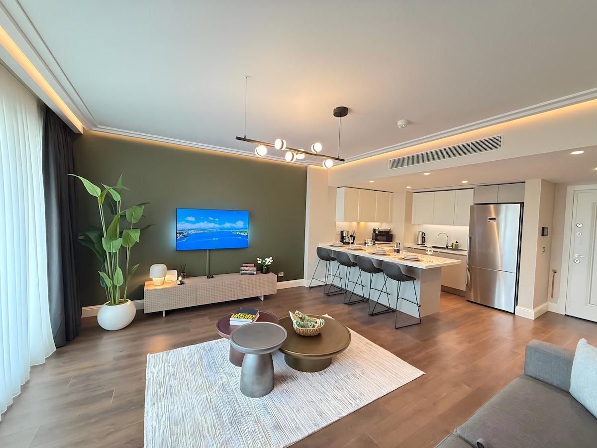 A modern living area displays a sleek TV mounted on a green accent wall. A light, neutral-toned sofa and coffee table are situated on a soft area rug. The open kitchen features contemporary appliances, and a tall plant adds a touch of greenery.