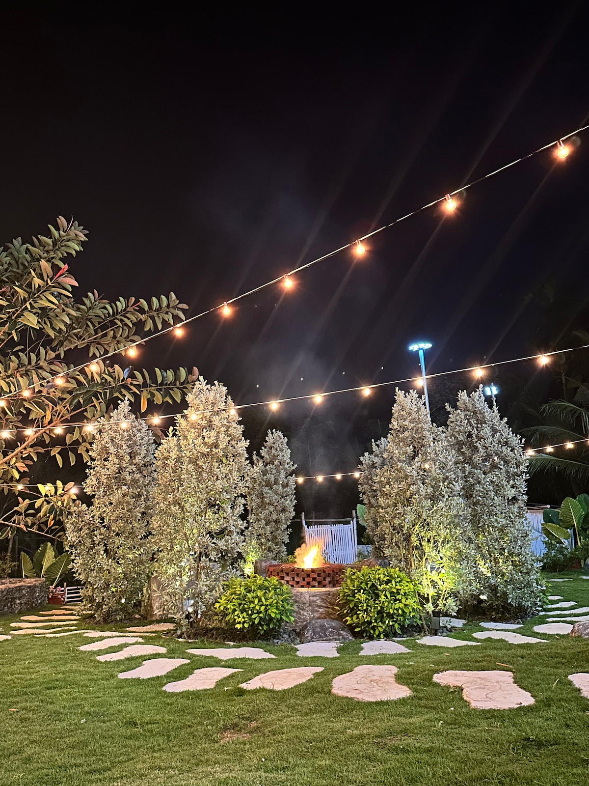 An inviting outdoor fire pit area is surrounded by neatly trimmed hedges and illuminated by string lights overhead. A winding stone path leads through a lush garden featuring greenery, with soft lighting highlighting the peaceful ambiance of the space at night.
