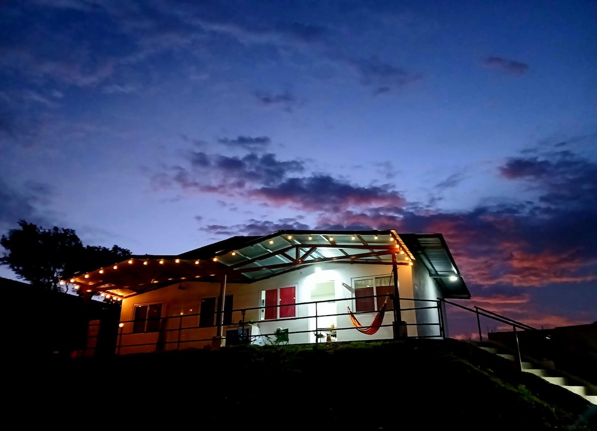 The exterior of a cozy home is illuminated by soft string lights during twilight. The structure features a metal roof and is surrounded by a grassy area. A hammock is visible, and a small seating arrangement can be seen by the entrance, with colorful clouds in the evening sky.