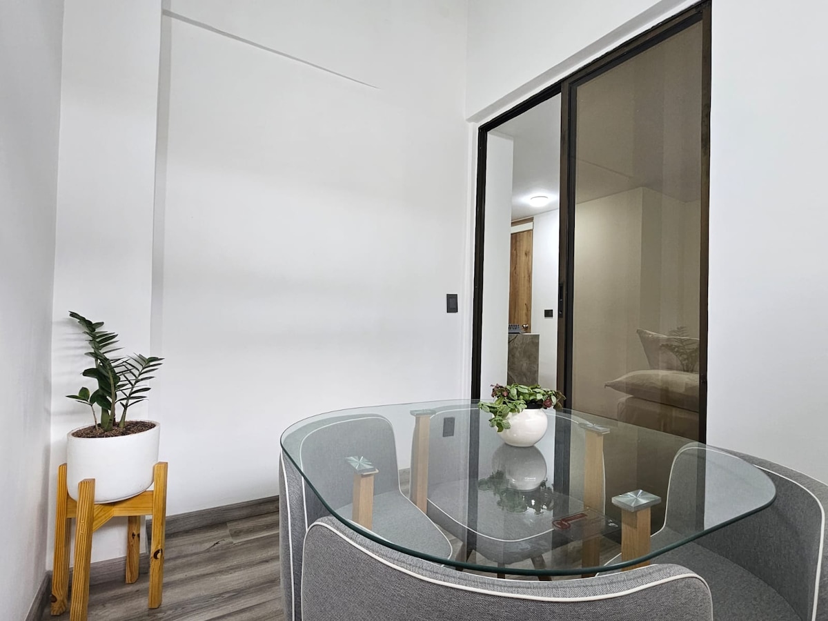 A round glass dining table with a minimalist design is surrounded by soft gray seating. A small potted plant adds a touch of greenery. Natural light filters through the glass sliding doors leading to an inner patio.