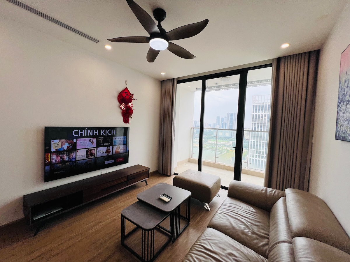 The spacious living area is furnished with a leather sofa and a coffee table, facing a large flat-screen TV mounted on the wall. Floor-to-ceiling windows provide natural light and showcase a view of the city skyline. A ceiling fan offers additional comfort.