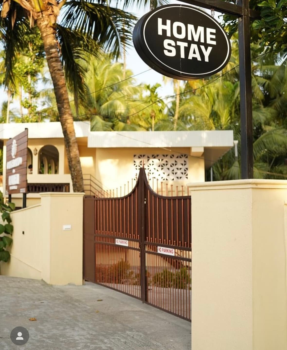 An inviting entrance features a decorative gated entry with a welcoming 'Home Stay' sign above. Lush palm trees provide greenery on either side, complementing the warm, earthy tones of the exterior walls. The path leading inside is paved, enhancing accessibility.