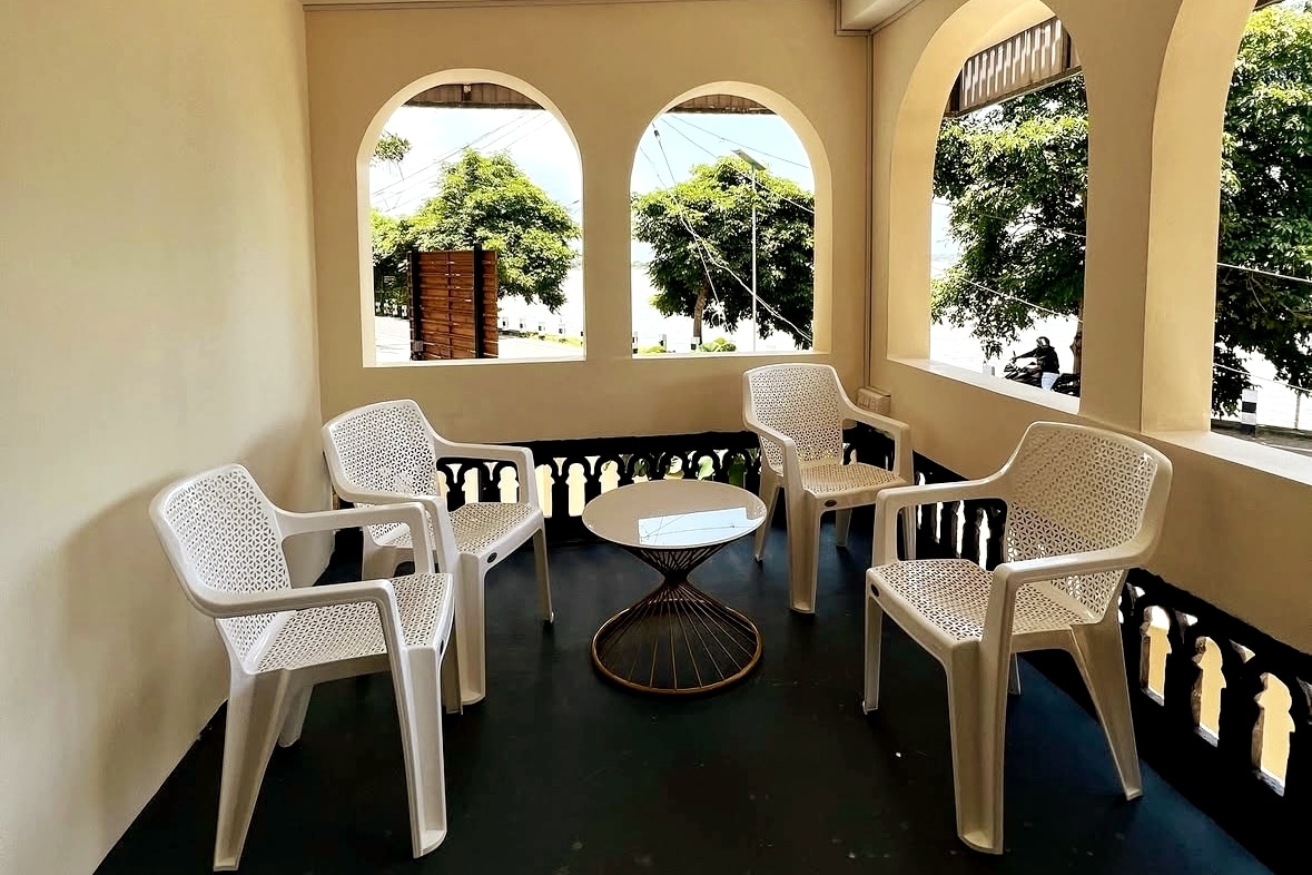 A shaded balcony area features four white plastic chairs arranged around a round table. The space is framed by arched openings, allowing views of greenery outside, providing a tranquil setting for relaxation.