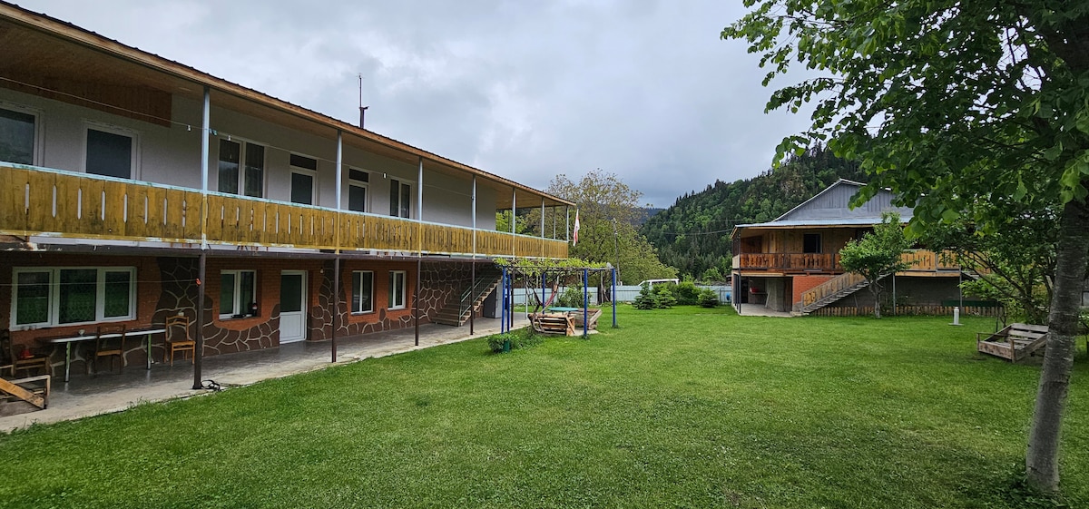 A spacious outdoor area is surrounded by two buildings, featuring a grassy lawn dotted with trees and seating arrangements. A covered porch extends from one building, while swings can be seen in a play area. Scenic mountain views provide a serene backdrop.