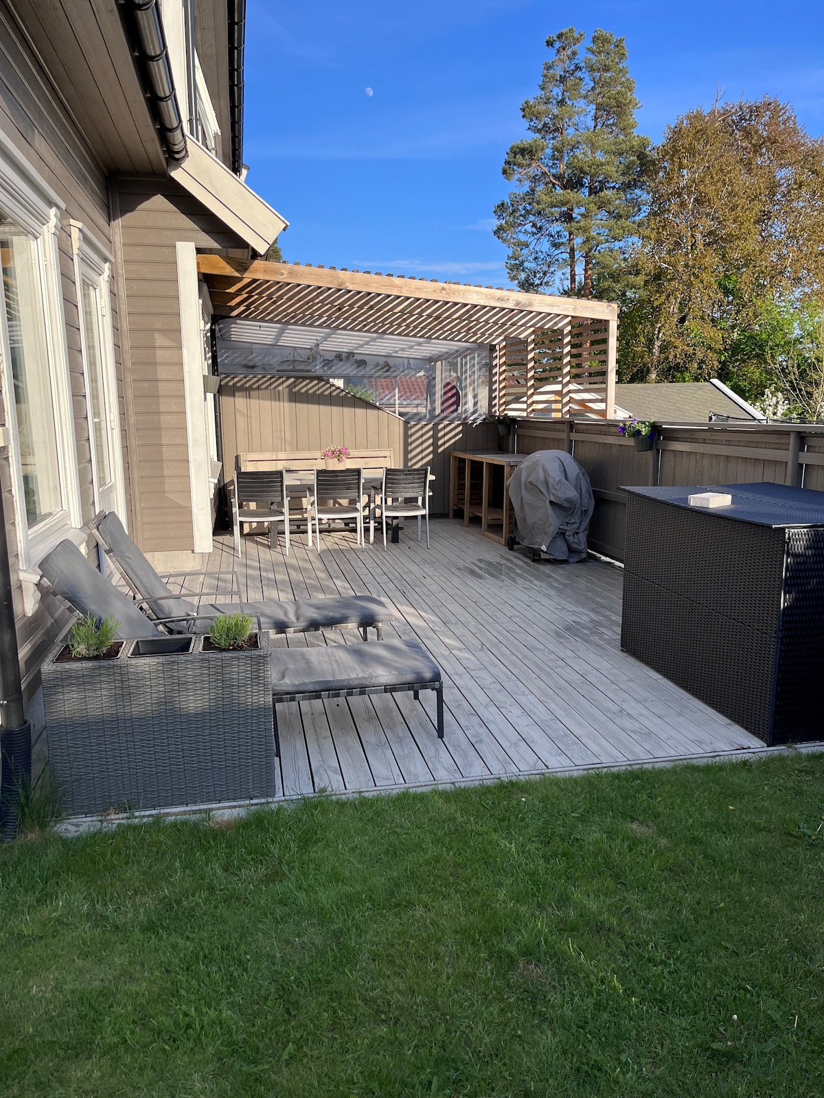 An outdoor terrace is presented, featuring a wooden deck and comfortable lounge chairs. A dining set with chairs is situated under a covered area. A barbecue grill is positioned on one side, while a dark storage unit is visible nearby. Lush green grass surrounds the space.
