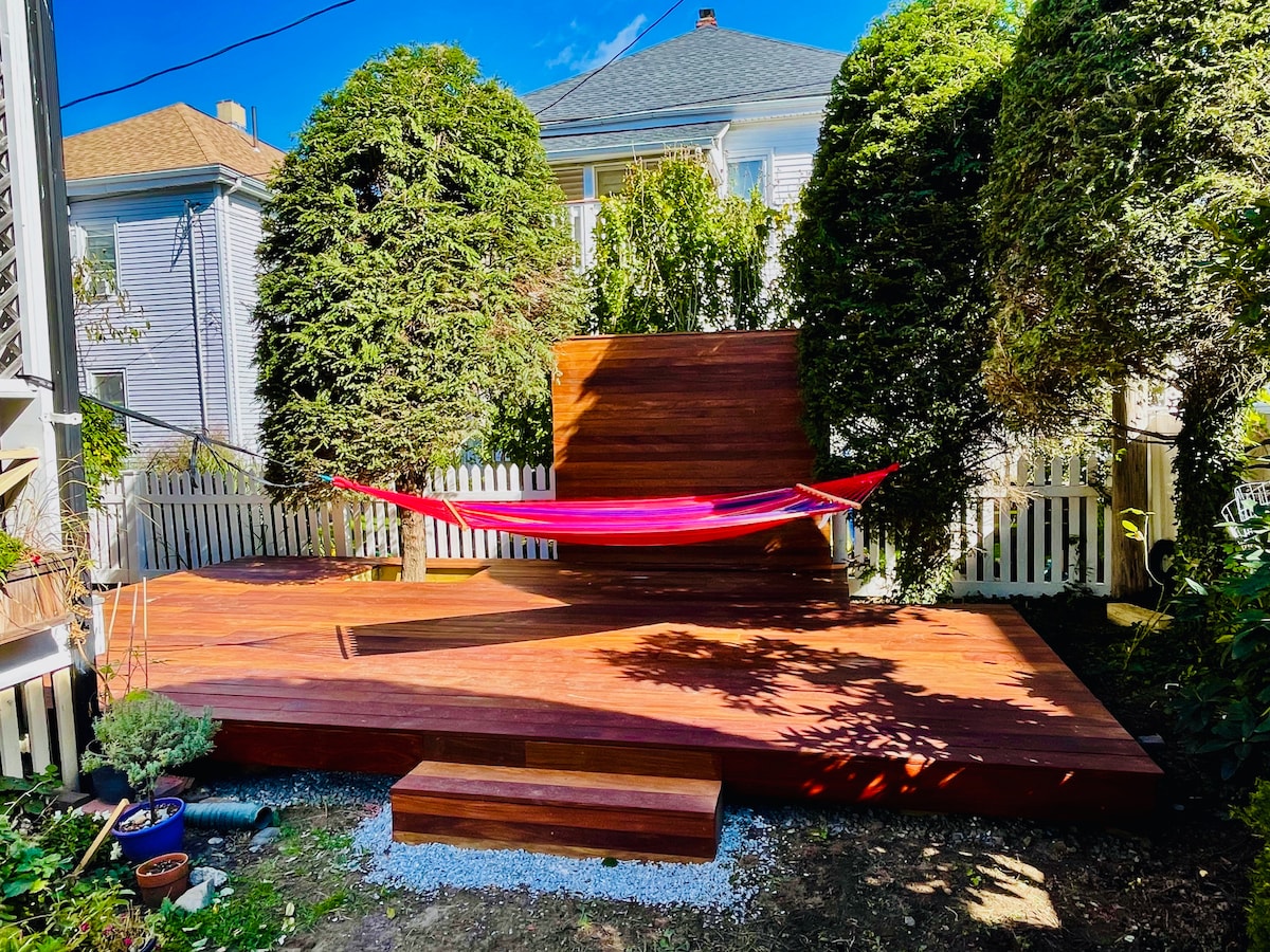 An outdoor deck area is bordered by lush greenery, featuring a vibrant hammock suspended between two trees. The deck is crafted from warm wood, with steps leading down to a landscaped yard. A white picket fence encloses the space, providing a sense of privacy.