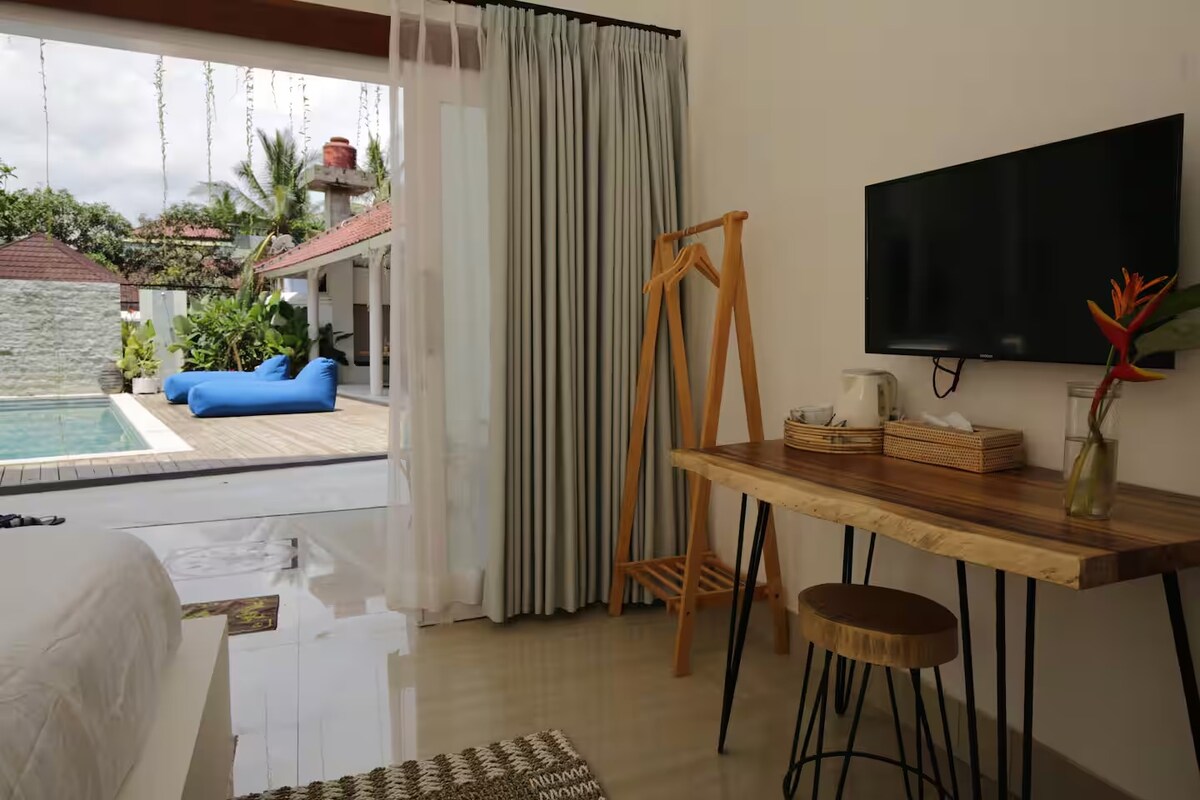 A bright room features a minimalist wooden desk and chair, accompanied by a television mounted on the wall. Natural light fills the space through large windows with sheer curtains, revealing views of a pool and tropical garden just outside.