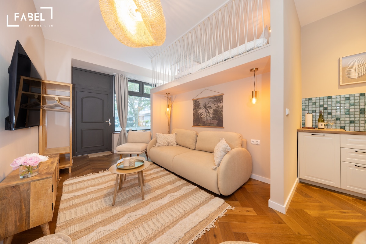 A spacious living area features a curved sofa, a glass coffee table, and wooden flooring. Natural light floods through the large window, while a flat-screen television is mounted on the wall. A decorative ceiling light hangs above, enhancing the modern design.