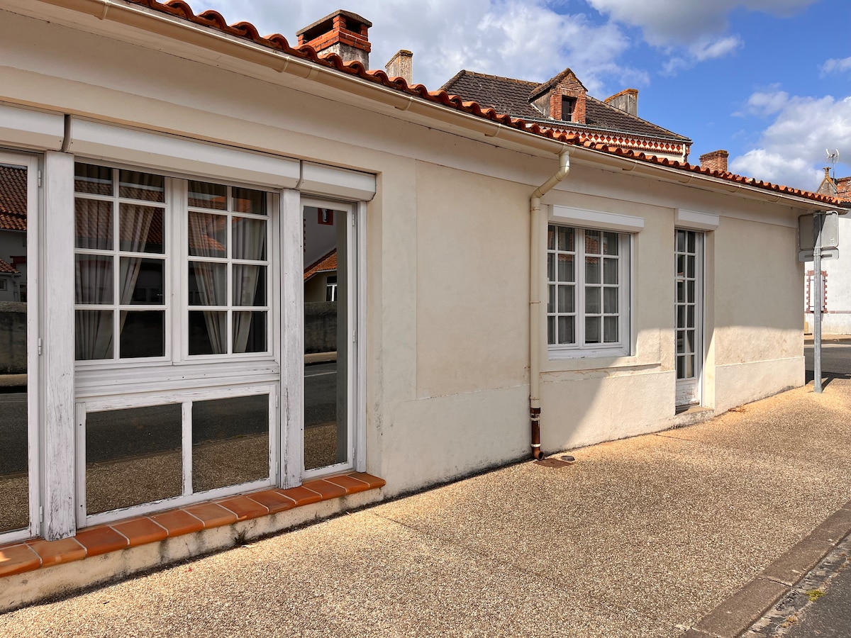 The exterior of the building features a charming, white facade with multiple window styles. Large glass windows allow natural light to enter, while the tiled area in front offers space for outdoor seating. The surroundings include a quiet street and nearby structures.