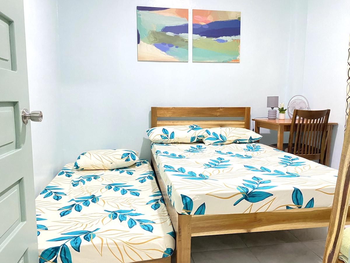 The bedroom features a wooden bed with a spacious mattress and a neatly arranged smaller mattress beside it, both adorned with light-colored linen with floral patterns. A small wooden desk and chair are present, complemented by a decorative plant.