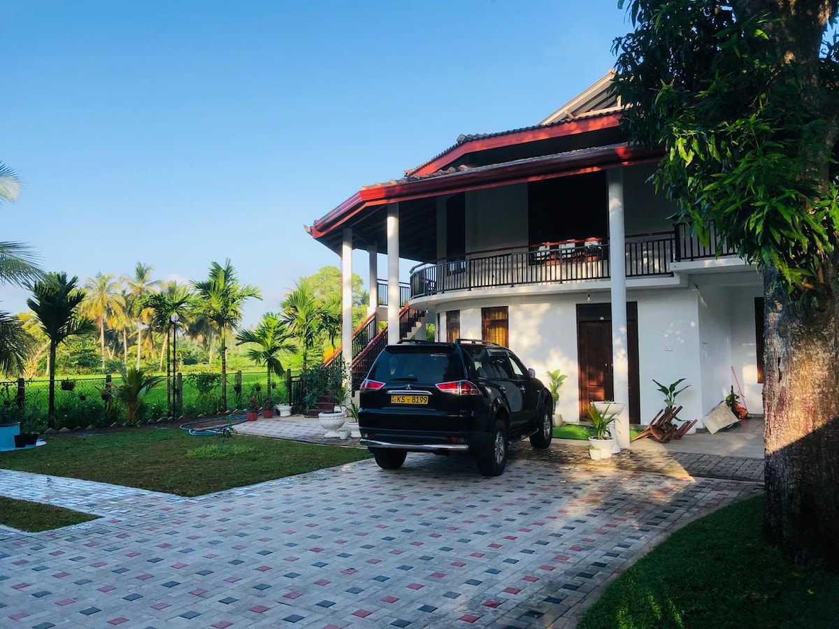 A two-story house features a covered porch with seating overlooking a lush garden. A black vehicle is parked on the paved driveway, bordered by well-maintained greenery. The expansive paddy fields are visible beyond the property, enhancing the serene atmosphere.