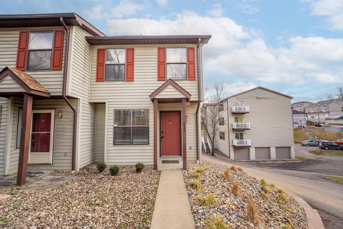 The entrance of a townhome is depicted, featuring a front pathway lined with gravel and small shrubs. Windows with red shutters are visible, along with a welcoming front door. Nearby, an additional building and parking area are shown in the background.
