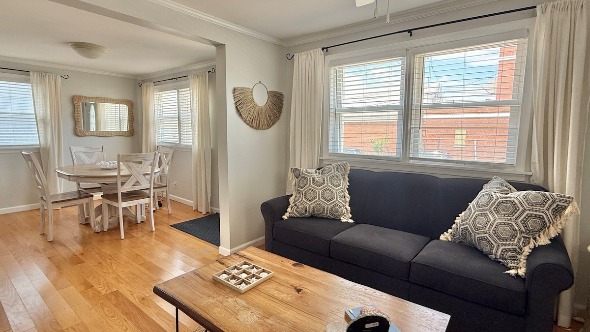 A living area features a dark sofa adorned with patterned throw pillows, while a wooden coffee table rests in front. A round dining table with white chairs is positioned nearby, illuminated by natural light from windows dressed in light, sheer curtains.