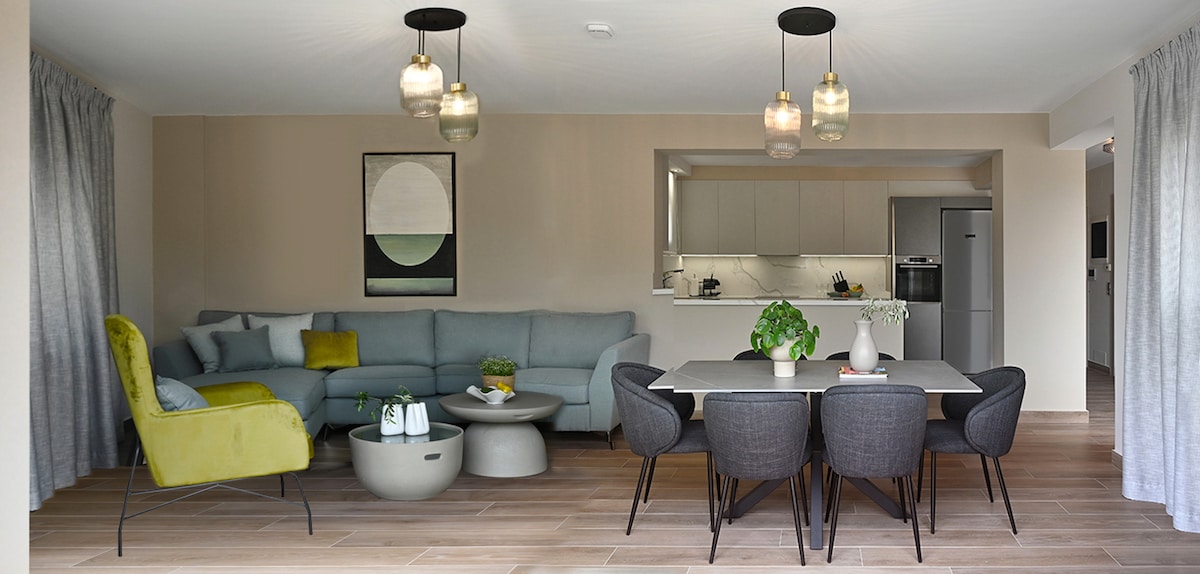 An open living space is presented with a light-colored sofa and a green accent chair. A round coffee table is positioned in the center, while a dining table is set for six nearby. Soft light is provided by modern pendant lamps above, enhancing a calming atmosphere.