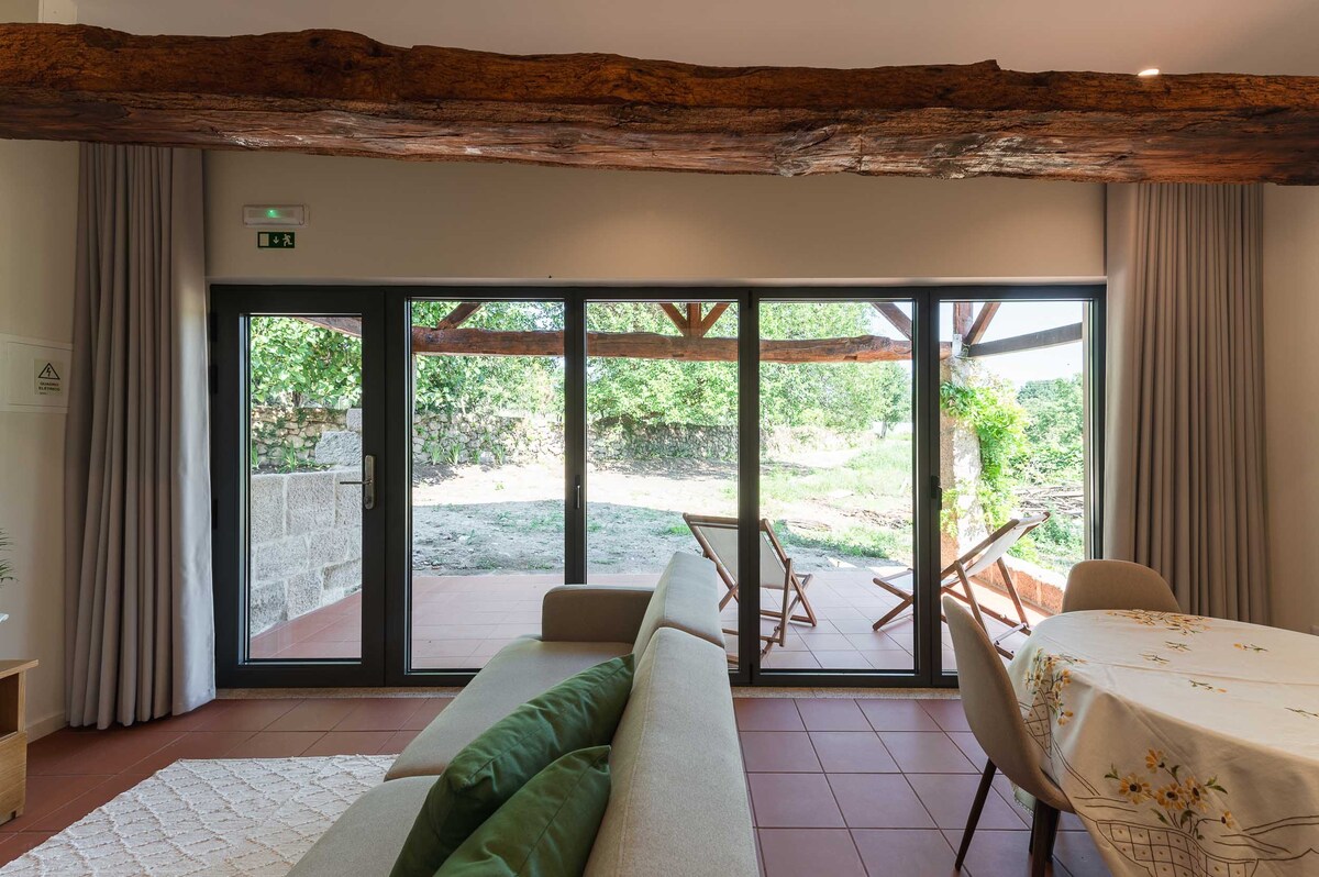 A spacious living area features a large glass door that opens up to a serene outdoor space. A beige sofa with green pillows is arranged neatly, while light-colored curtains frame the windows, allowing natural light to fill the room. Exposed wooden beams add an element of rustic charm.