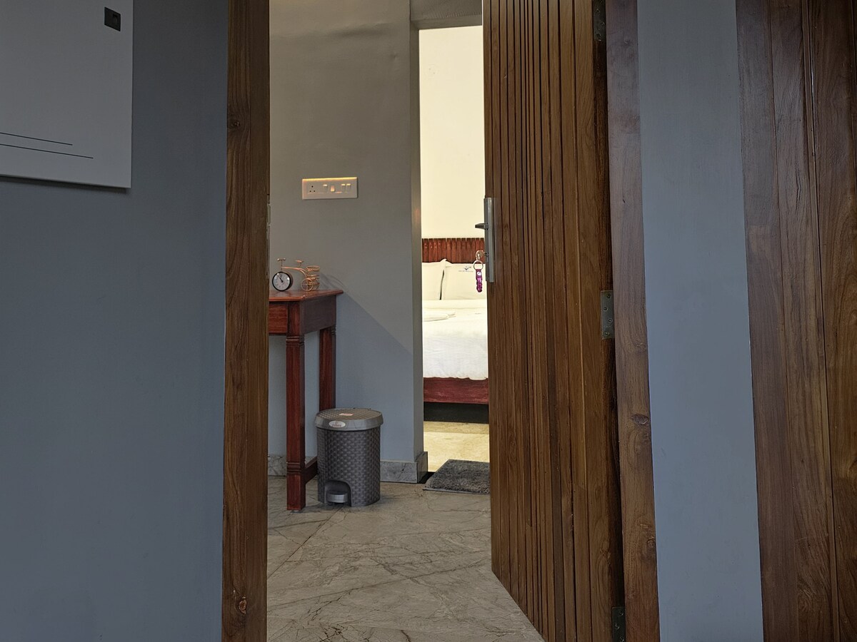A wooden door is partially opened, revealing a glimpse of a bedroom in the background. The room features a spacious bed and is accentuated by a small wooden table and a waste bin on the floor, enhancing the inviting ambiance of the interior.