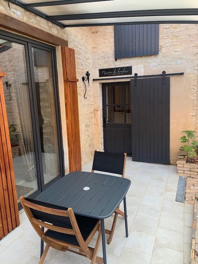 A serene outdoor patio features a black dining table with two wooden chairs. The space is enclosed by stone walls and framed by large glass doors that allow natural light to enter. A wooden door adds to the rustic charm of the area.