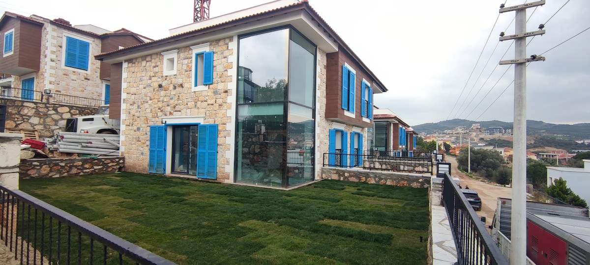 A modern villa with a stone facade is displayed, featuring large glass windows and blue shutters. A well-maintained green lawn surrounds the property, enhancing the inviting outdoor space. The surrounding area includes other homes and distant hills, contributing to a relaxed atmosphere.