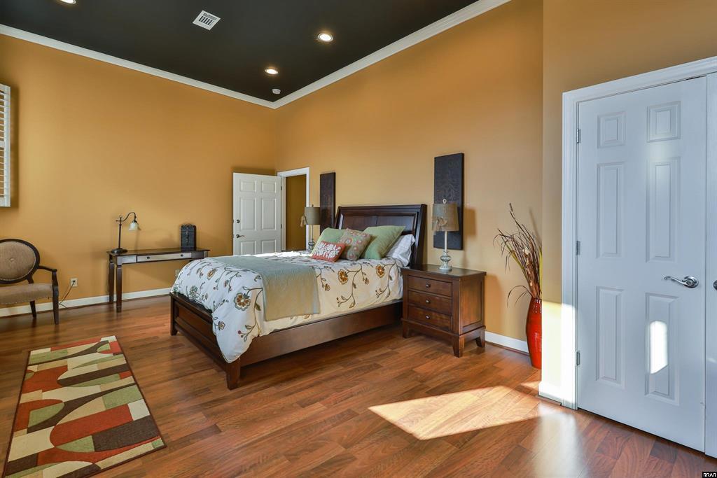 A spacious bedroom features warm golden walls with a cozy bed dressed in soft linens. A wooden dresser and a sleek desk add functional elements. Natural light floods the room through the door, illuminating the rich wooden flooring and decorative accents.