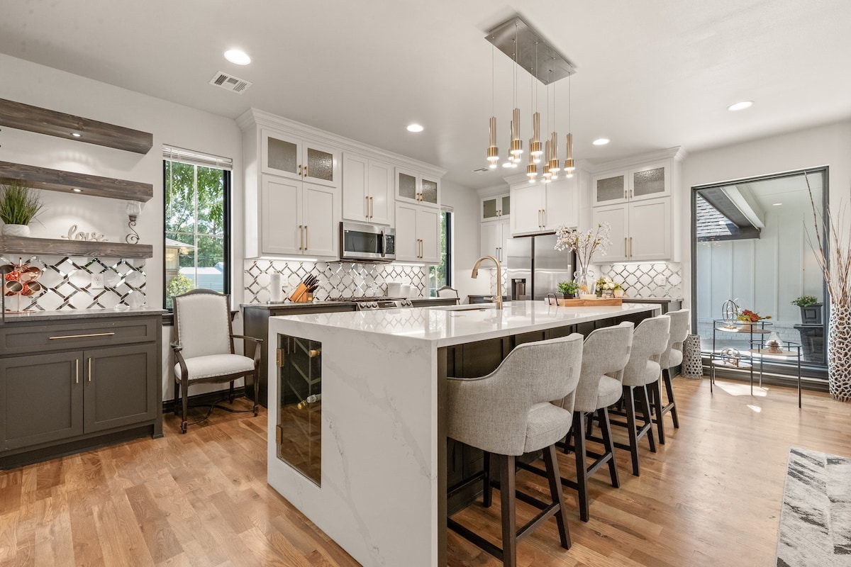 A modern kitchen features an expansive island with seating for several. Light wood flooring complements the neutral cabinetry. Stylish light fixtures hang above, illuminating the space. Large windows provide natural light, and a door opens to an outdoor area with seating options.