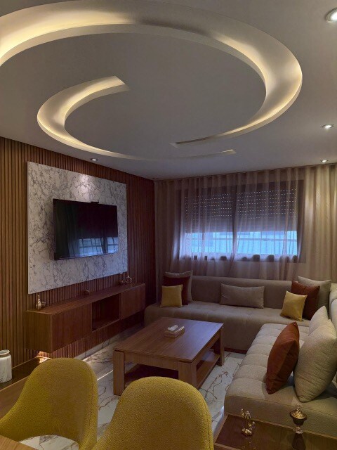 A modern living area features a large television mounted on a decorative marble wall. Soft lighting from the ceiling fixture creates a calming ambiance. A comfortable sectional sofa, adorned with various cushions, surrounds a wooden coffee table on a stylish patterned rug.