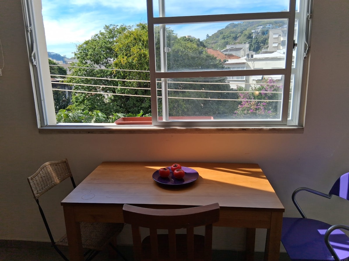 A simple wooden table is positioned next to a window, providing a view of greenery outside. Two chairs accompany the table, one made of wood and the other in a vibrant color. Natural light illuminates the scene, highlighting a round plate with three decorative items.