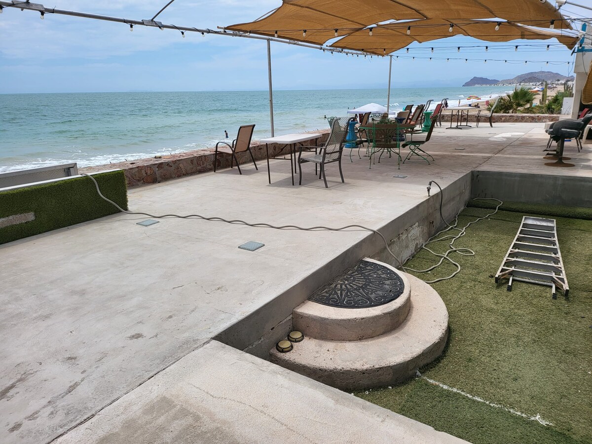The spacious patio is shown with a view of the Sea of Cortez, featuring a mix of seating options, including chairs and lounge areas. A grassy area is partially visible, along with a pool edge and a round mat. Light cords are placed along the patio.