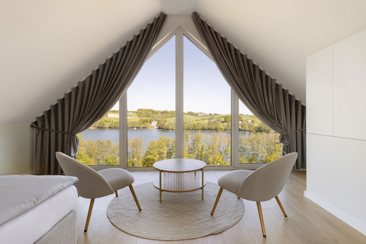 A spacious room features large triangular windows dressed in soft, dark drapes, offering views of lush greenery and a calm lake. Two comfortable armchairs are positioned beside a circular coffee table, providing an inviting space for relaxation.