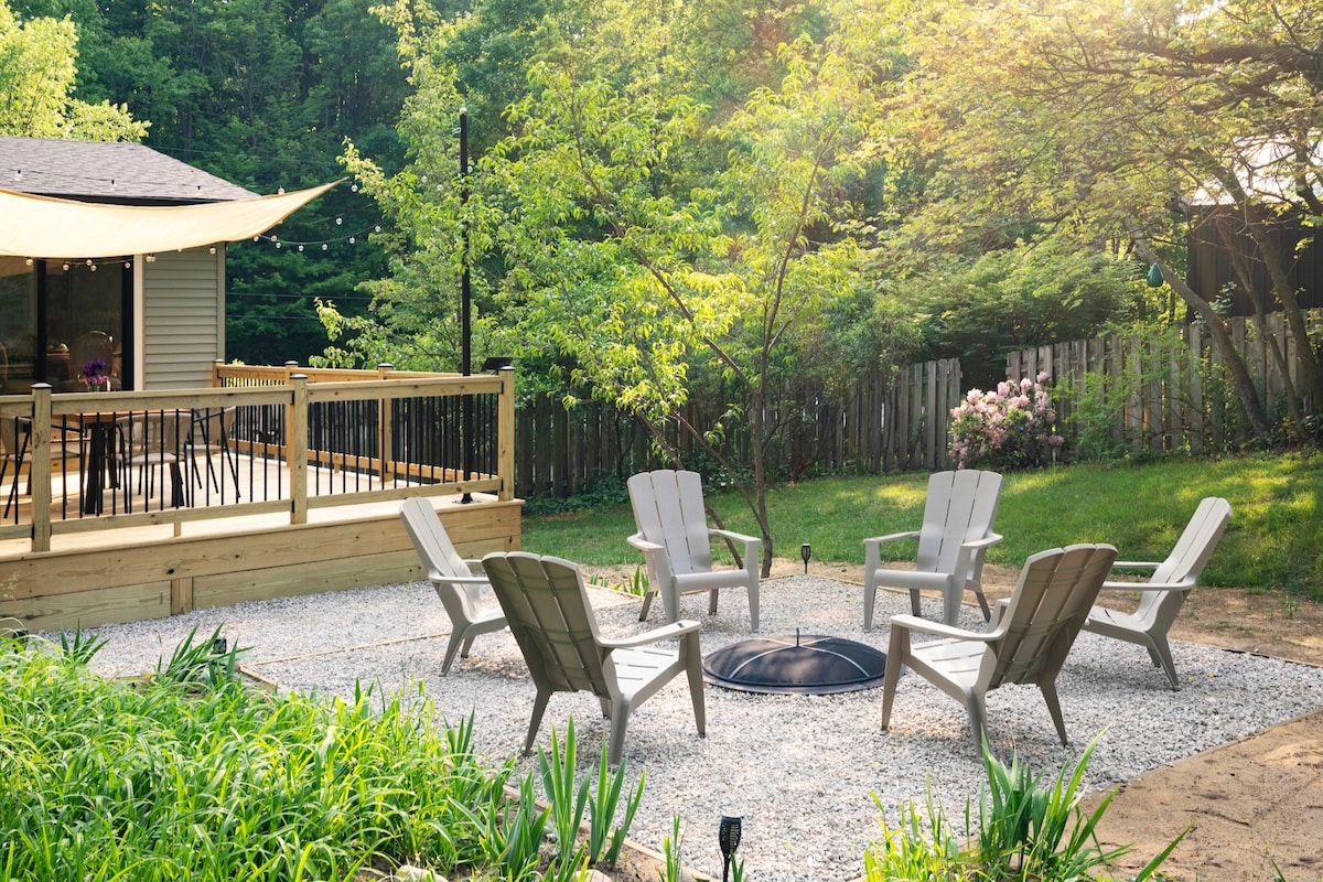 An outdoor seating area is framed by greenery, featuring five light-colored adirondack chairs arranged around a central fire pit on a gravel surface. A wooden deck is visible in the background, complemented by sunlight filtering through the trees.