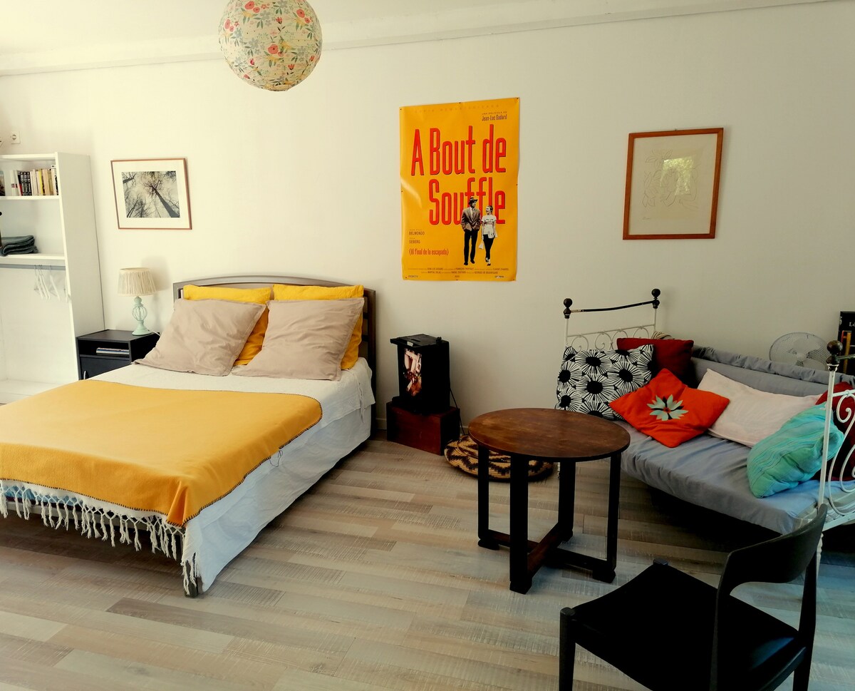 A well-lit sleeping area is featured, showcasing a spacious bed with a yellow and white coverlet. A small round table sits between the bed and a comfortable seating area. Colorful artwork, including a movie poster, adorns the walls, enhancing the space's character.