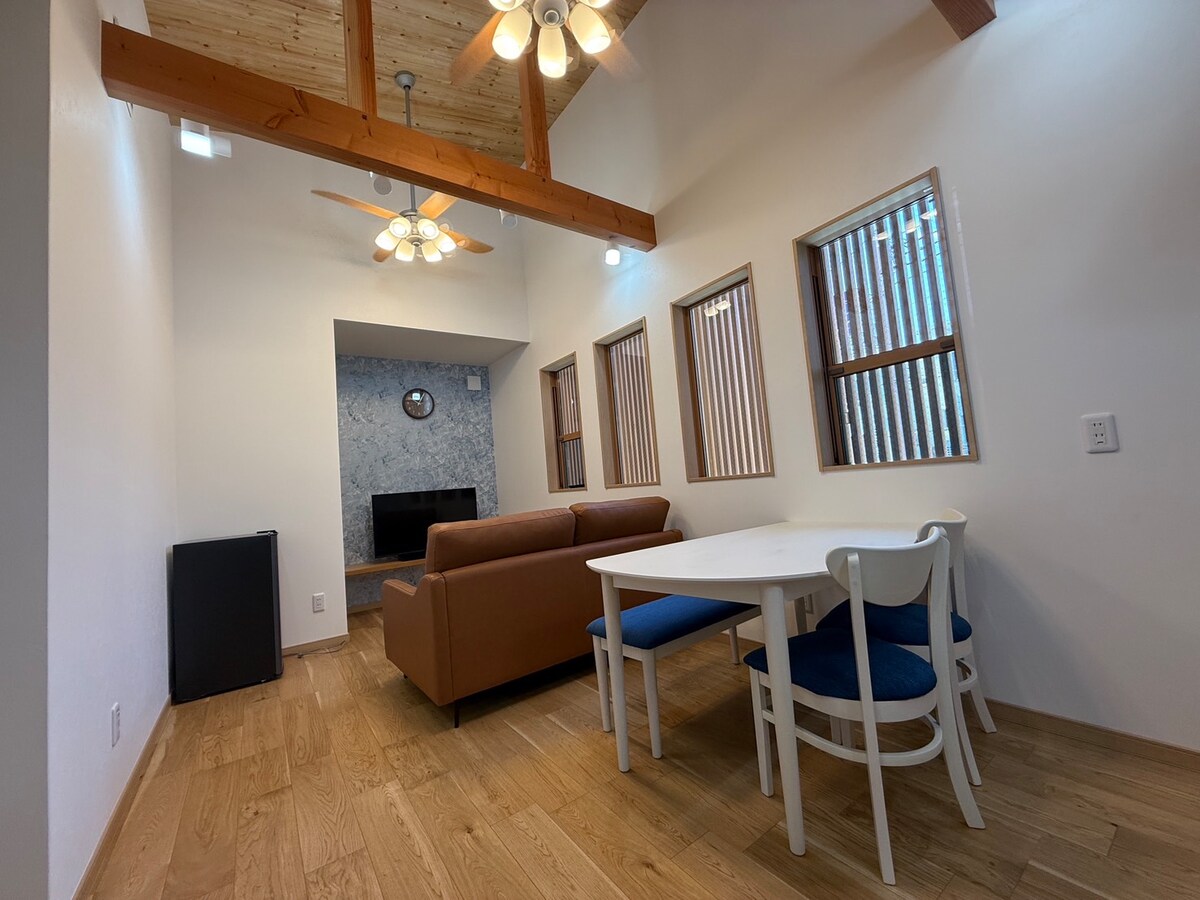A well-lit living area features a brown sofa facing a wall-mounted television. A dining table with white and blue chairs is situated next to the sofa. Natural light filters through multiple windows, highlighting the wooden ceiling beams and warm wooden flooring.