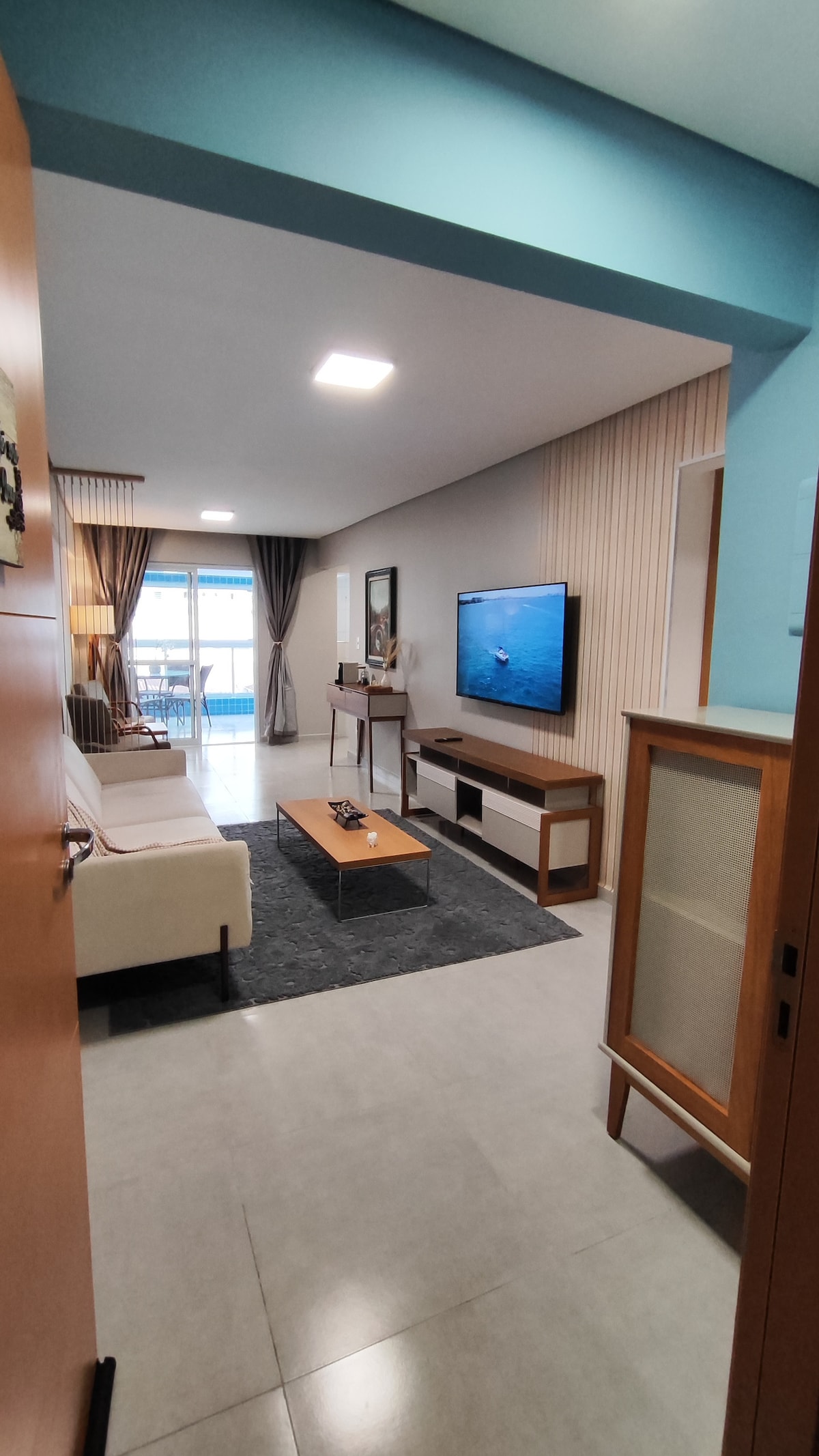 The living area is designed with a neutral color palette, featuring a comfortable sofa and a wooden coffee table. A television is mounted on a console against the wall. Natural light filters in through large glass doors that lead to a balcony, framed by curtains.