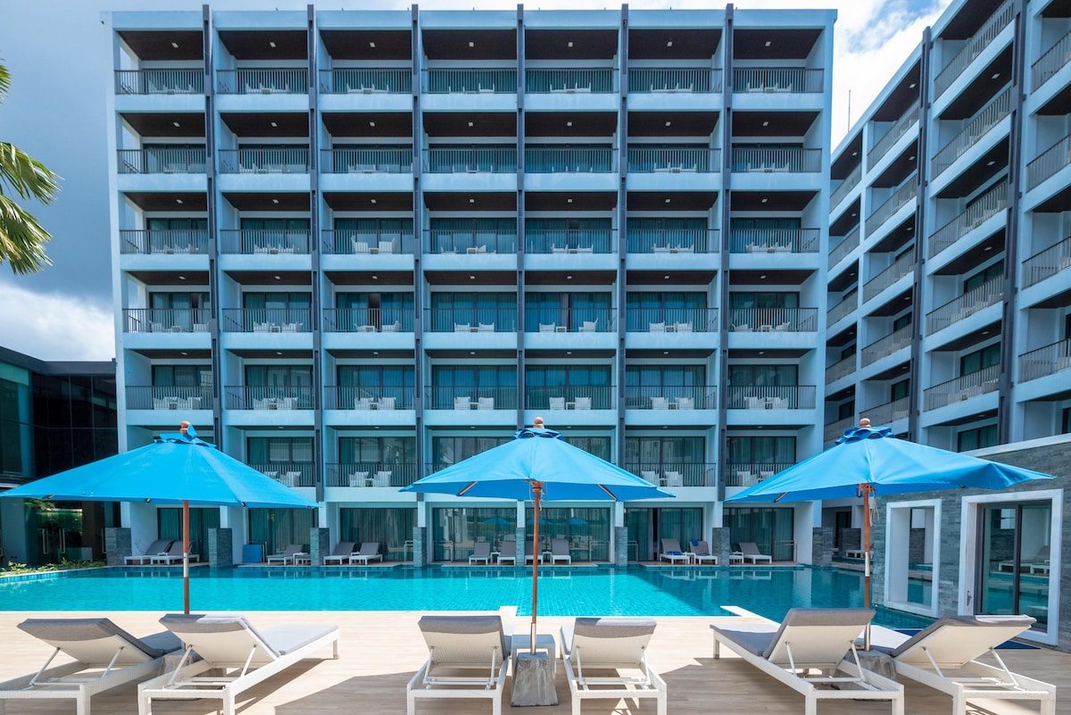 A large outdoor pool is surrounded by spacious loungers and blue umbrellas, offering a relaxing atmosphere. The modern apartment building rises in the background, with multiple balconies visible on each floor, reflecting a blend of comfort and contemporary design.