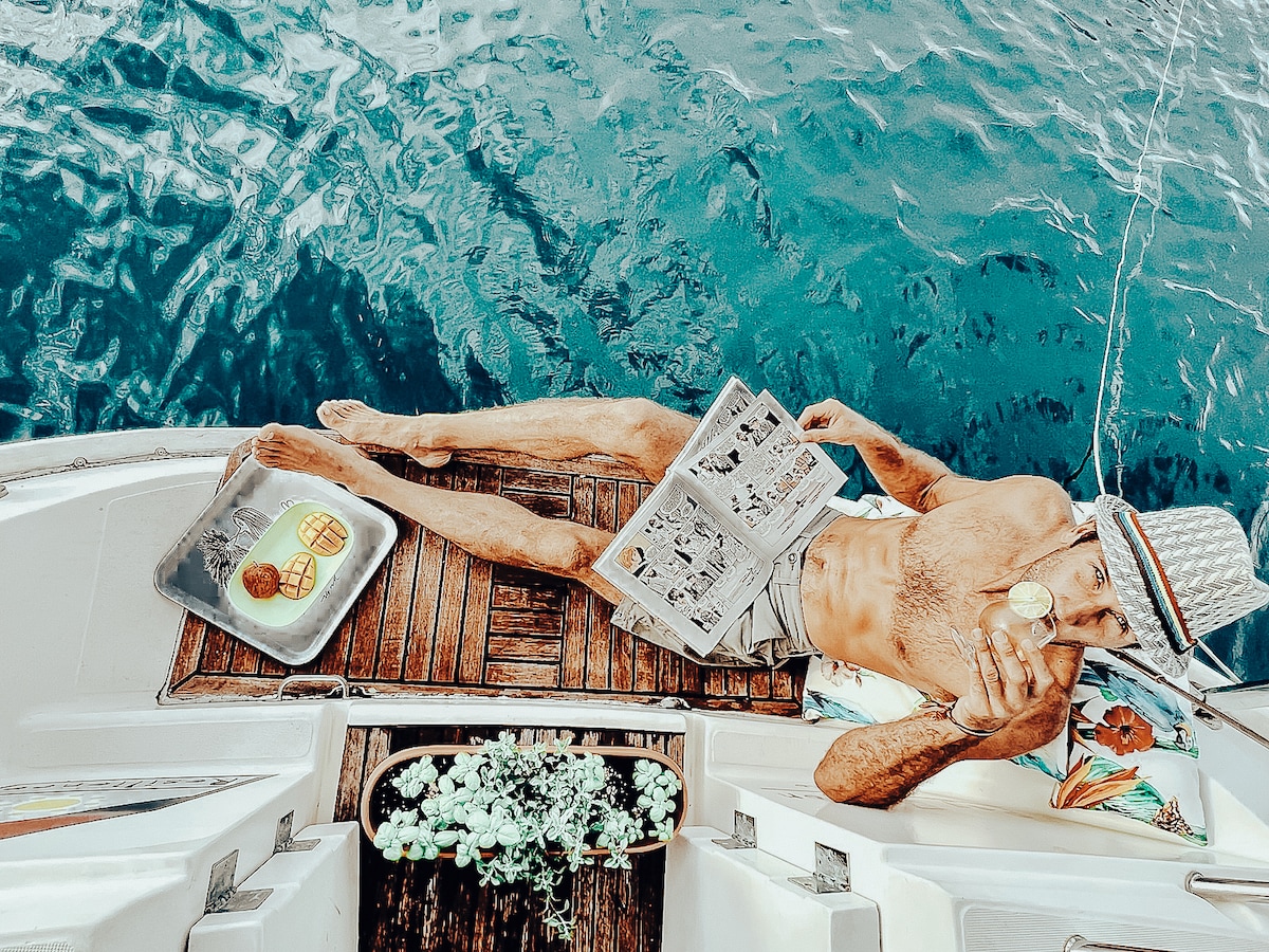 A person relaxes on the deck of a boat, enjoying a drink while reading a newspaper. A tray with a light meal is placed nearby. The deep blue water reflects sunlight, creating a tranquil atmosphere. Plants and colorful fabric add a touch of vibrancy to the setting.