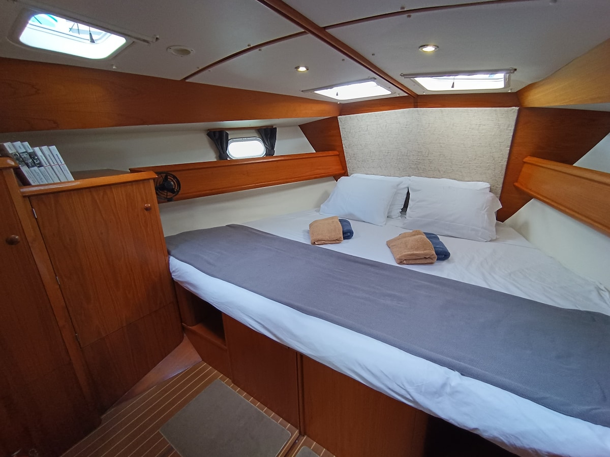 The spacious cabin features a queensize bed with crisp white linens, accented by two towels placed artfully on the bed. Wooden cabinetry provides ample storage, while natural light filters through hatches above, enhancing the comfortable atmosphere.