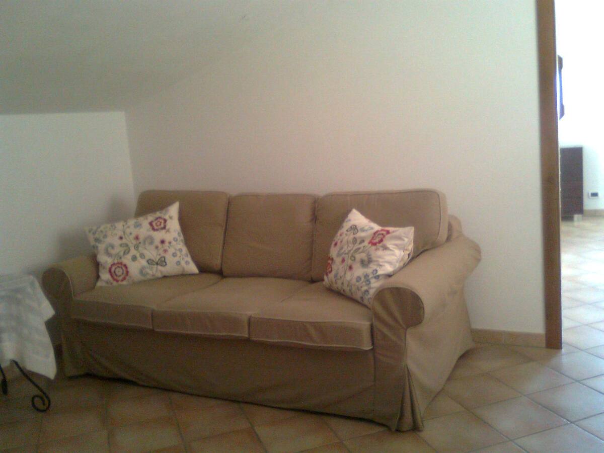 A cozy living area is highlighted by a neutral-toned sofa adorned with decorative throw pillows featuring floral patterns. The floor is covered with warm tile, and light walls create an inviting ambience. A doorway leads to another space, suggesting accessibility.