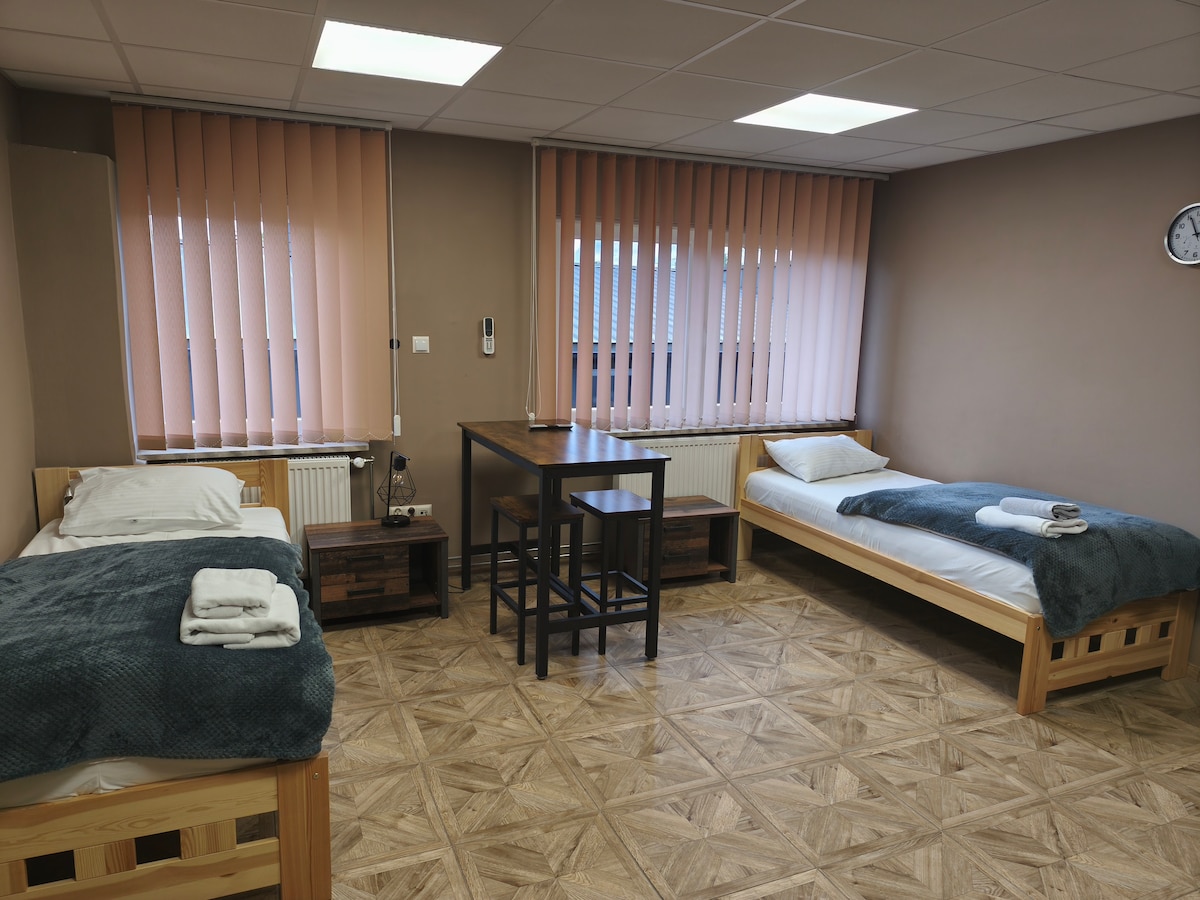 A spacious room is furnished with two single beds, each topped with fresh linens and towels. A small dining table surrounded by four chairs is positioned in the center. Light filters through vertical blinds, while a clock is mounted on the wall.