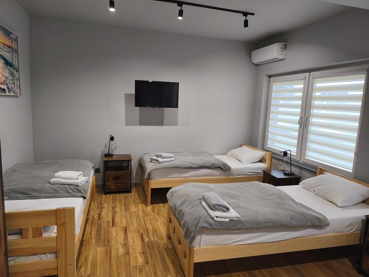 A modern guest room features three spacious beds arranged thoughtfully against light grey walls. Each bed is neatly made with soft linens and towels. A flat-screen television is mounted on the wall, and natural light filters through the partially open window coverings.