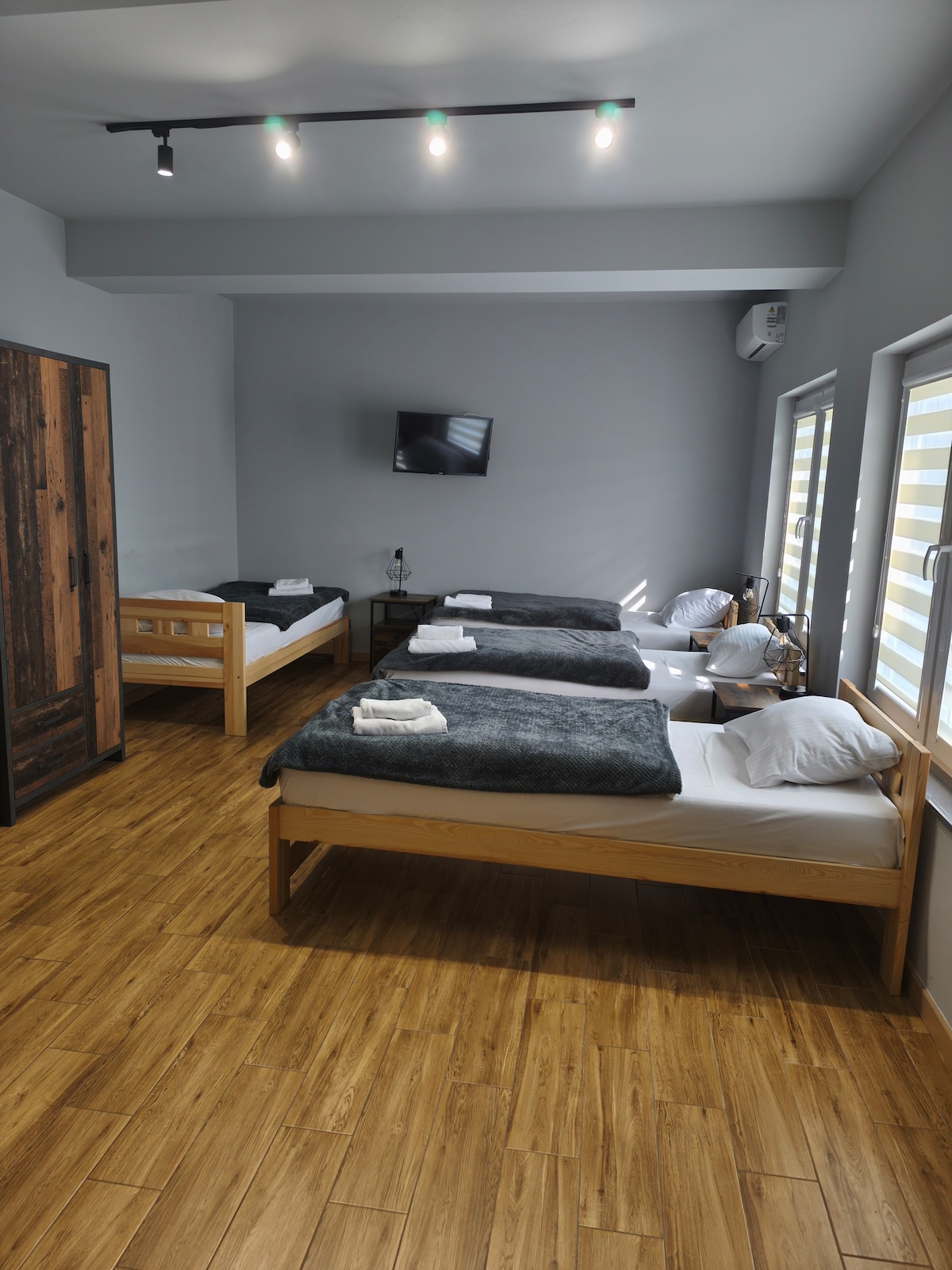 A bright sleeping area is furnished with multiple wooden beds, each topped with gray blankets and white pillows. A television is mounted on the wall, and natural light enters through the partially covered windows, enhancing the space's modern design.