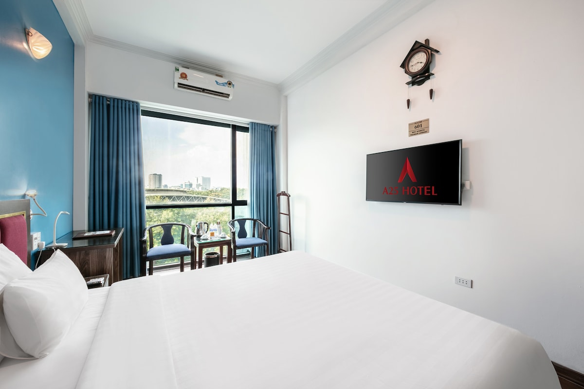 A bright and airy hotel room features a spacious bed with crisp white linens. A flat-screen television is mounted on the wall, while a small seating area with two chairs and a table overlooks the view through large windows, framed by blue curtains.