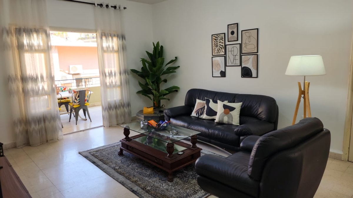 A spacious living area is furnished with dark leather sofas positioned around a glass coffee table. Natural light pours in from the balcony doors, illuminating the room. Tropical plants are arranged thoughtfully, and framed artwork adorns the walls, adding a touch of style.