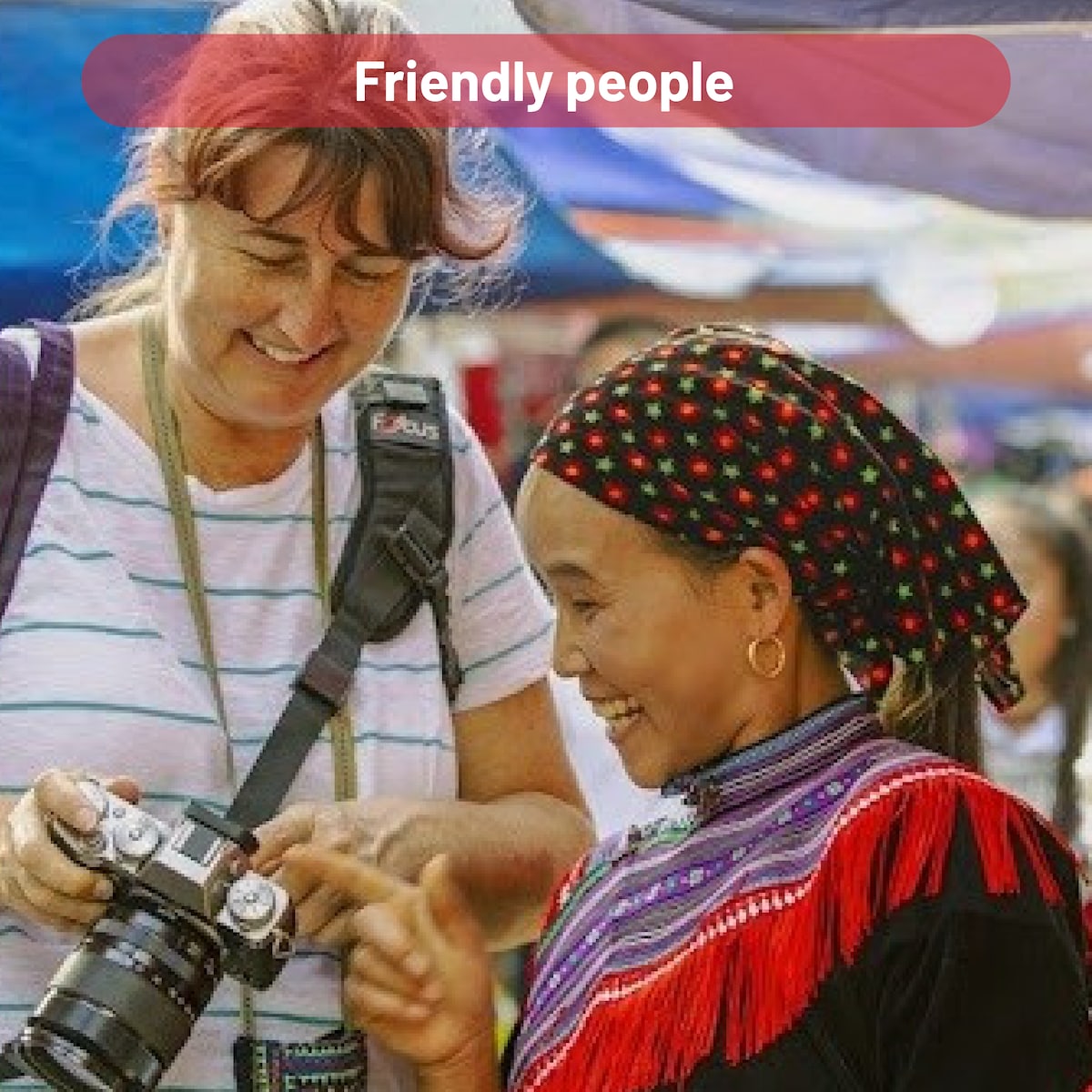 Two individuals are interacting in a vibrant outdoor market, showcasing diverse traditional attire. One person is holding a camera, while the other is dressed in colorful garments and intricate accessories. The bustling market atmosphere is enhanced by colorful fabrics and various goods in the background.