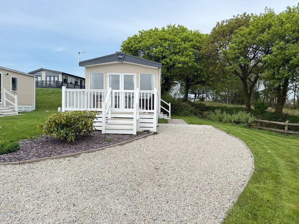 A charming cabin is positioned among greenery, featuring a white exterior with a porch and large windows. A gravel path leads to the entrance, bordered by manicured landscaping. Trees provide natural shade to the surrounding area, enhancing the tranquil setting.