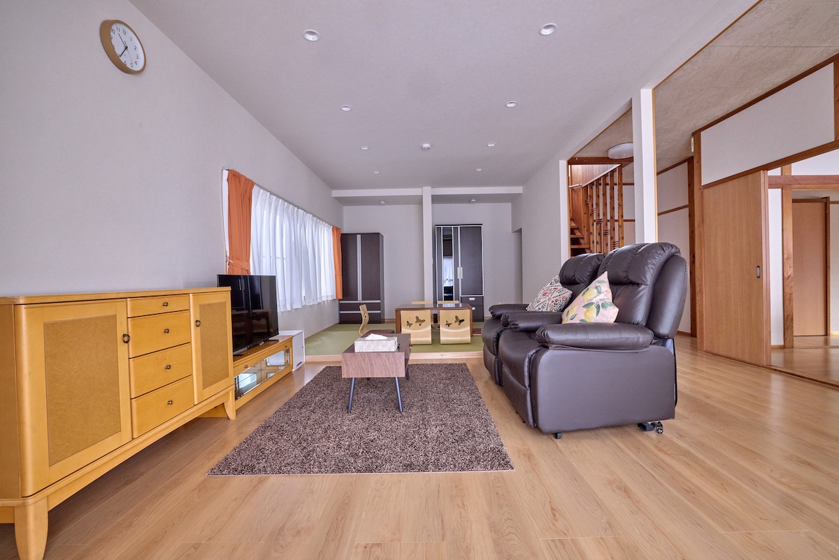 A spacious living area features a large area rug on wooden flooring, with comfortable leather seating positioned around a central coffee table. A television is mounted on an elegant cabinet, while large windows allow natural light to fill the space, creating a bright and airy environment.