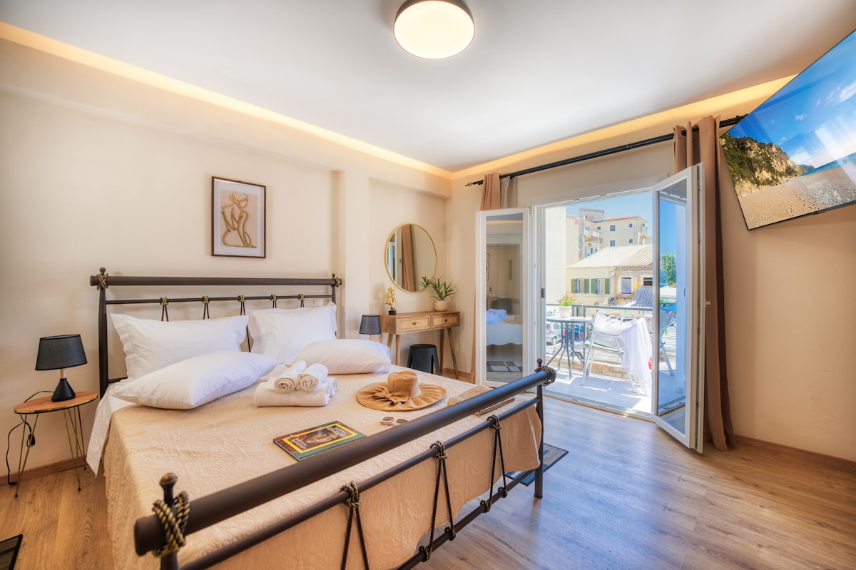 A bright sleeping area features a spacious bed with soft linens and decorative pillows. Large glass doors lead to a private balcony, allowing natural light to fill the room. A round mirror and a small wooden desk create a functional and stylish environment.