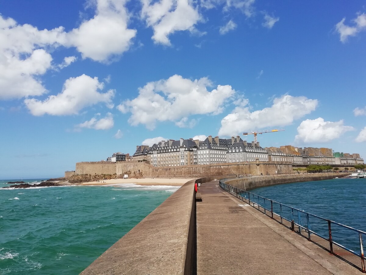 A stone jetty extends toward a coastal town, with distinct buildings featuring sloped roofs lining the shoreline. The blue water contrasts with the sandy beach, while clouds pass over a clear sky, enhancing the view of the historic fortifications in the background.