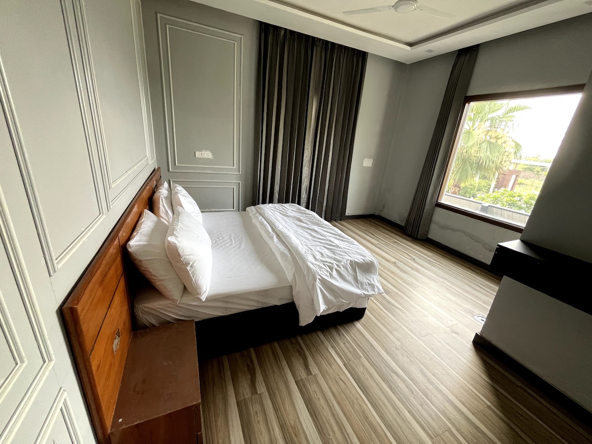 A spacious sleeping area features a neatly made bed with crisp white linens. Natural light enters through a large window framed by dark curtains, highlighting the room's wooden flooring and minimalist decor, including a wooden nightstand and a window showcasing views of the garden.