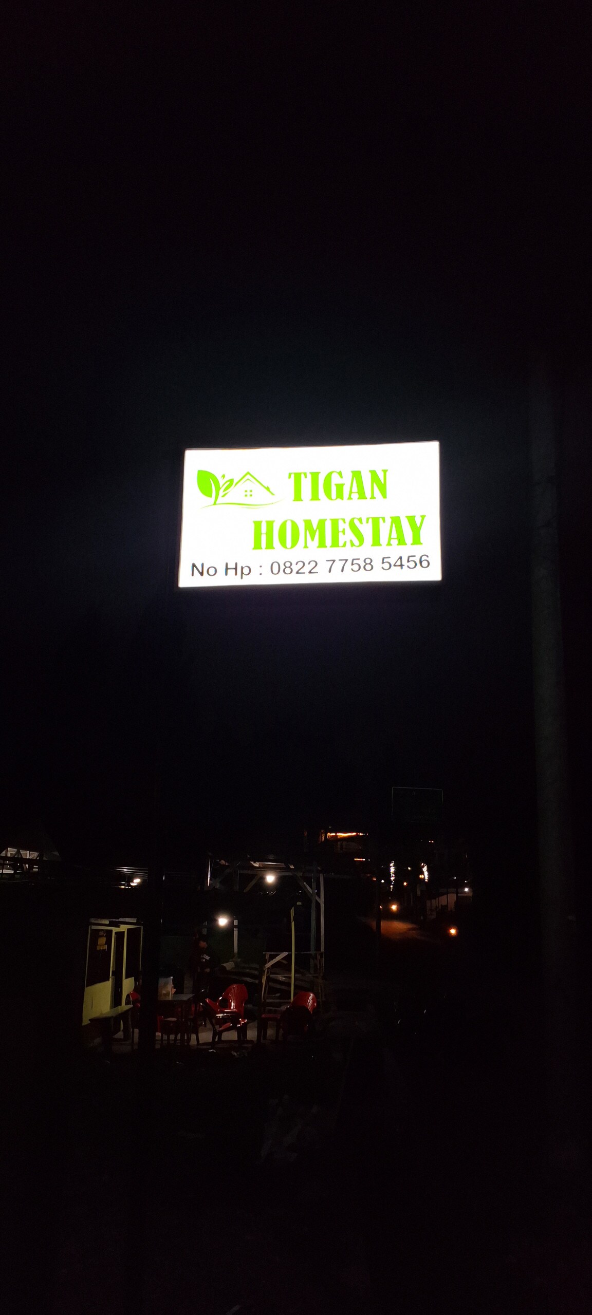 A brightly illuminated sign displaying 'Tigan Homestay' stands prominently against a dark night backdrop. Nearby lights hint at additional structures and furniture, creating a visible sense of a welcoming environment.
