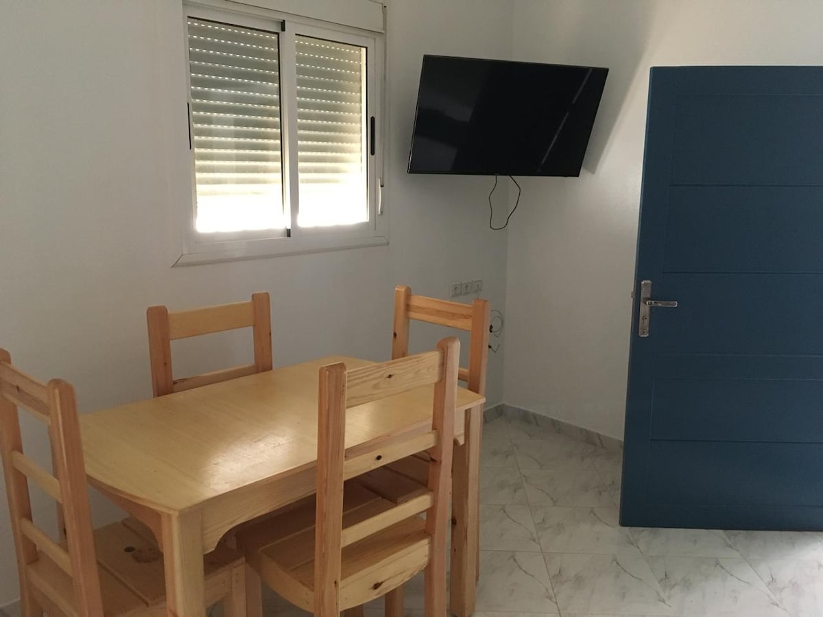 A simple dining area features a light wooden table surrounded by four matching chairs. A flat-screen television is mounted on the wall, and a window with horizontal blinds allows natural light to enter the room. The space is completed by a blue door.